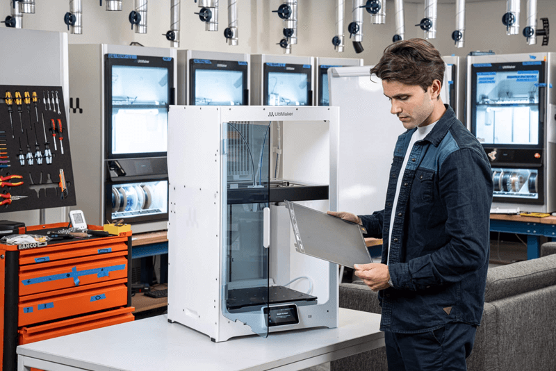man håller i en byggplatta framför en vit 3d skrivare